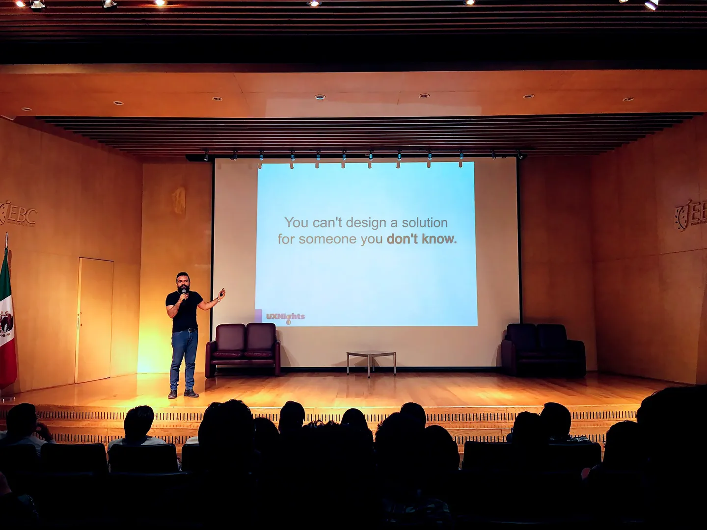 Sadok Cervantes on stage in a warm wooden auditorium, a slide behind him